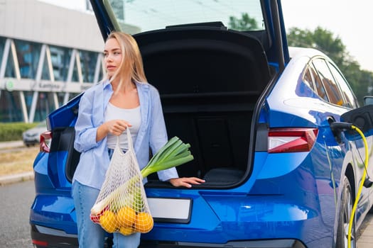 Young woman with groceries stands by the open trunk of a blue electric car in an urban setting.