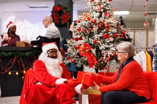Older client sitting with santa claus in shop, receiving amazing christmas gift from employee next to festive ornaments. African american worker spreading holiday cheer in shopping mall.