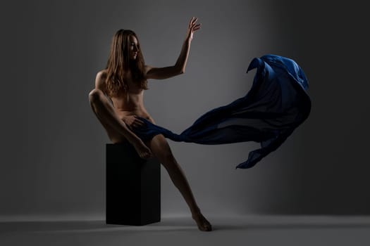 Slim pretty longhaired brunette sitting on a cube with fluttering blue cloth against gray background