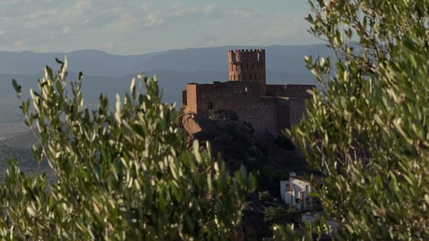 A slow-motion shot captures an old castle on rugged rocks at sunset, with moving tree branches in the foreground and side light casting dramatic shadows.