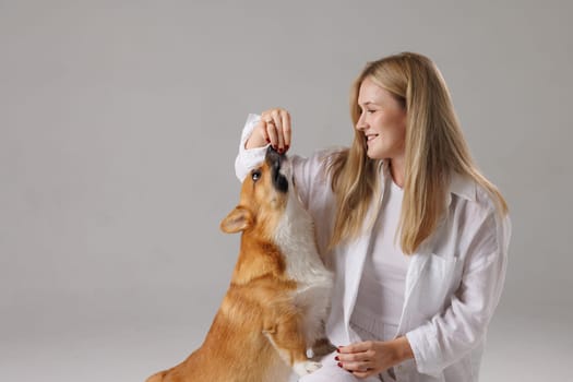 A woman in a white outfit is feeding a treat to a corgi dog in a studio setting.