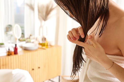 A woman with wet hair is inspecting the ends of her hair while standing in a bright, airy bathroom. She is wrapped in a towel, and there are beauty products on a wooden dresser in the background.