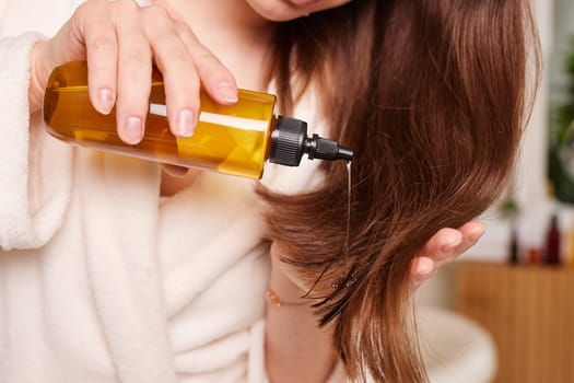 Woman applying oil on her long shiny hair against bathroom background, hair health care