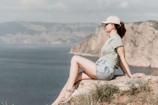 A woman wearing a green T-shirt, denim shorts, and a cap sits on a cliff, facing a vast blue sea and a misty mountain range in the distance.