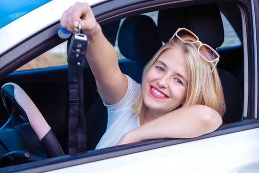 A young woman is sitting in the car and holding her new key, smiling with joy as she looks out a side window.