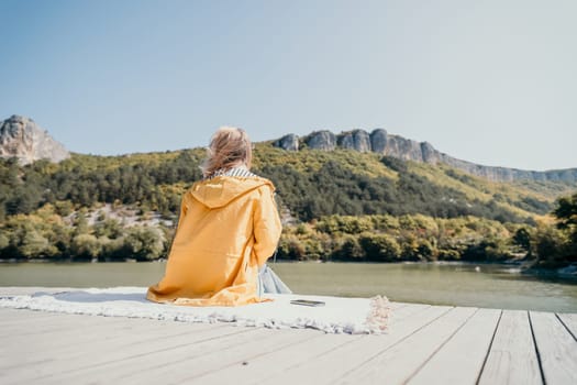 A woman in a yellow jacket sits on a wooden dock, gazing towards a mountain range by a calm lake.