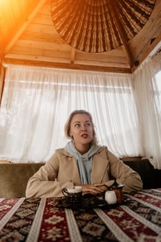 A young woman in a light blue hoodie and beige coat sits at a table in a cozy cafe, looking thoughtfully towards the left, a cup of coffee in front of her.