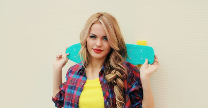 Portrait of stylish young blonde woman posing with skateboard on city street against white wall background