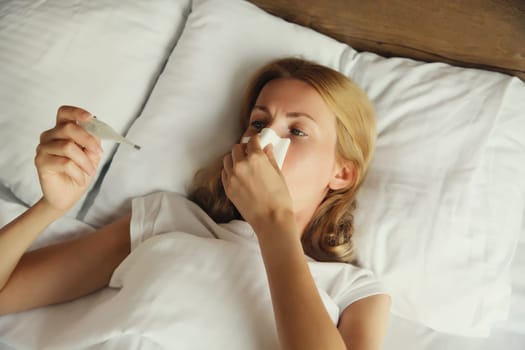 Sick upset woman sneezing blow nose using tissue and measuring checking body temperature looking at thermometer on the bed in bedroom at home