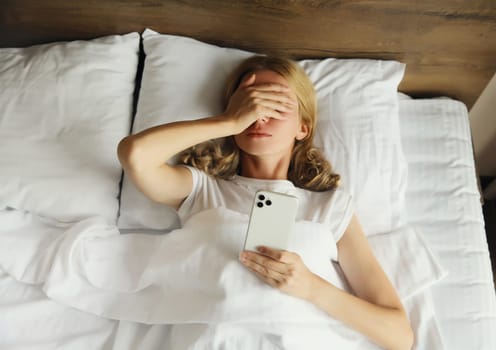 Sad upset young woman with phone, crying girl holding smartphone lying on the bed in bedroom at home