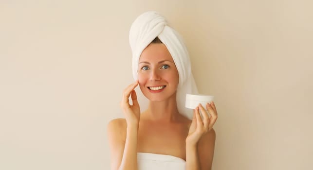 Natural beauty portrait of happy smiling young caucasian woman touches her clean skin applying face cream while drying wet hair with white bath towel on her head