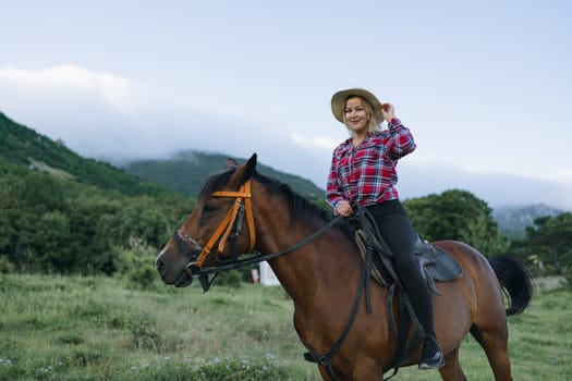 A woman is riding a horse in a field. The woman is wearing a plaid shirt and a straw hat. The horse is brown and is standing in a grassy field. The sky is blue