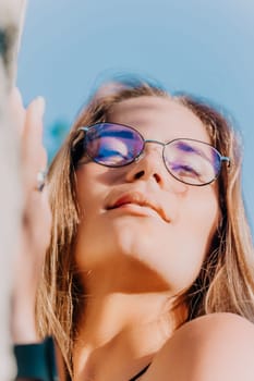 A woman with long blonde hair wearing purple-tinted glasses looks up at the sun.