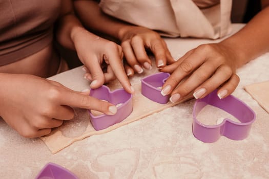 Two hands are using purple heart-shaped cookie cutters to cut dough. The dough is on a wooden surface, and the hands are in focus.