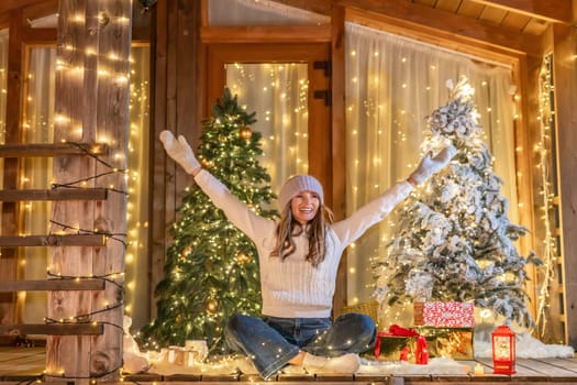 A woman sits on a porch, surrounded by Christmas trees and lights, with her arms raised in the air. She is smiling and appears to be happy.