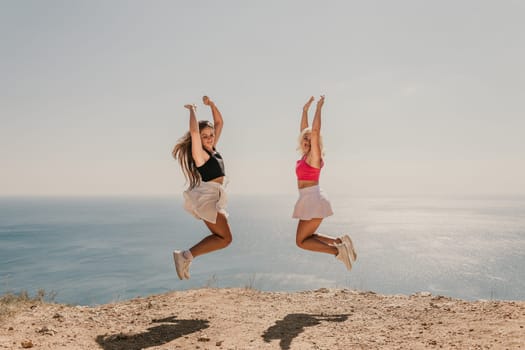 Two women are jumping in the air on a beach. The sky is blue and the ocean is in the background. Scene is joyful and energetic