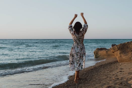 A woman in a floral dress stands on a sandy beach, arms raised in the air, facing the ocean. The water is choppy and the sky is blue.
