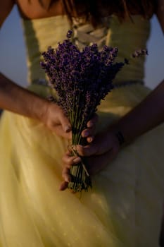 Woman poses in lavender field at sunset. Happy woman in yellow dress holds lavender bouquet. Aromatherapy concept, lavender oil, photo session in lavender.