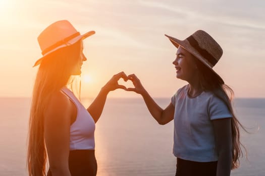 Two women wearing straw hats stand side by side, facing each other and making a heart shape with their hands as the sun sets over the ocean.