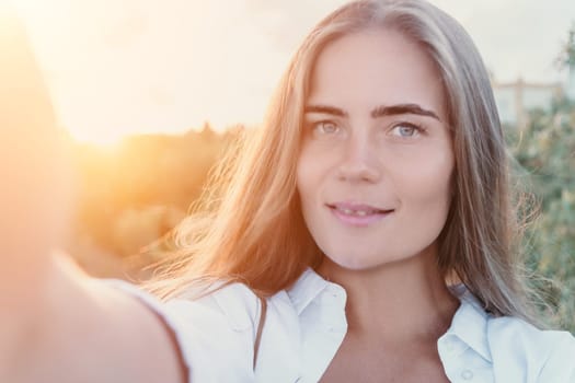 A young woman with long blonde hair takes a selfie with the sun behind her. She is smiling at the camera. She is wearing a white shirt.