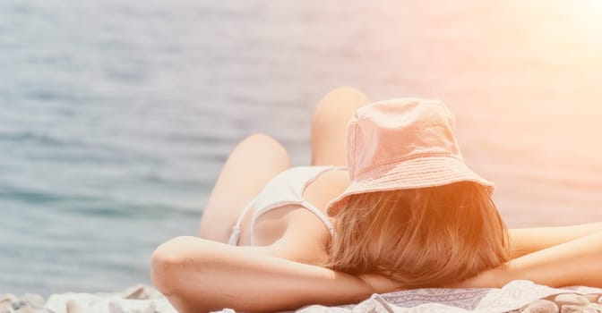 A woman sunbathing on a rocky beach, wearing a white swimsuit and a hat, with the sun shining brightly.