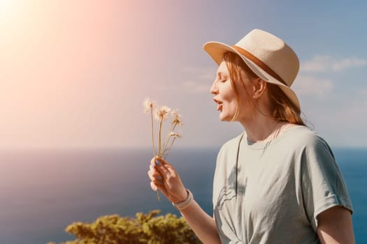 A young woman in a hat and a light green shirt blows on dandelion seeds while looking up at the sky.
