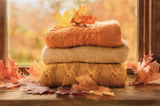 Stacked wool autumn sweaters on wooden background with leaves near the window