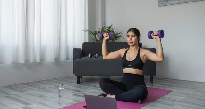 Young woman exercising in front of laptop Wear a sports bar outfit. Do yoga and lift dumbbells on the exercise mat..