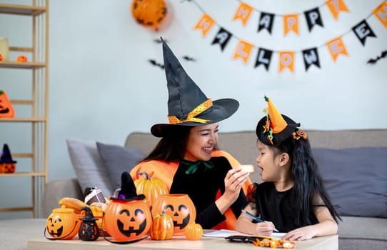 Playful young mother sitting on comfy couch in living room with cute little daughter tells spooky Halloween stories. Funny mom and girl kid have fun at home. Celebration and Autumn holiday concept.