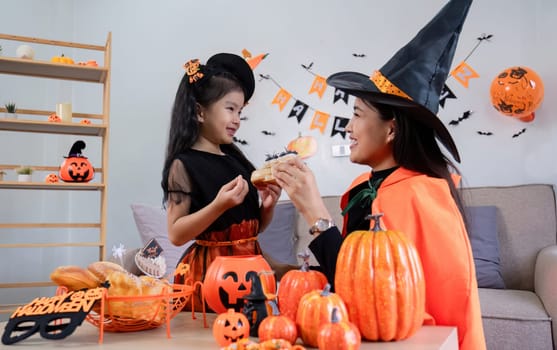 Playful young mother sitting on comfy couch in living room with cute little daughter tells spooky Halloween stories. Funny mom and girl kid have fun at home. Celebration and Autumn holiday concept.