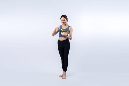 Young sporty Asian woman in sportswear holding salad bowl fill with vibrant of fruit and vegetable. Natural youthful and fit body lifestyle with balance nutrition on isolated background. Vigorous