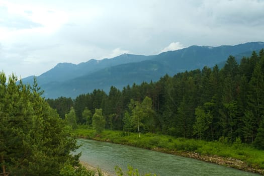 Amidst the mountains, a river flows gently through vibrant greenery, while clouds loom overhead, creating a peaceful atmosphere that invites contemplation and connection with nature.