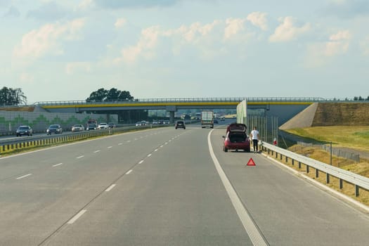 Gliwice, Poland - June 15, 2023: A man stands beside a red car with its trunk open on the side of a highway, placing a warning triangle on the road. The landscape features fields under a clear blue sky.