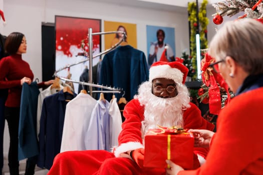 Senior person sitting with santa in store, accepting beautiful christmas present from worker next to festive decorations. African american adult spreading holiday joy in shopping center.