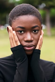 young tender african american female with short hair covering face with hands while looking at camera outdoors on summer day
