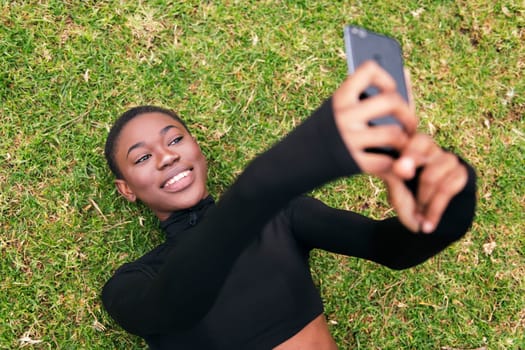 young african american woman with short hair smiling happy taking selfie photo on mobile phone lying on the grass in a park