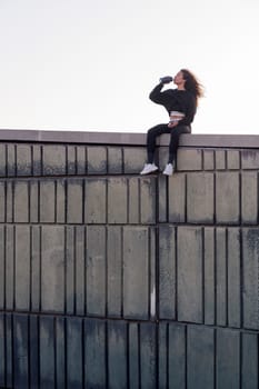 thirsty female athlete in sports clothes drinking beverage from bottle while resting on building roof after workout, concept of urban sport and healthy lifestyle