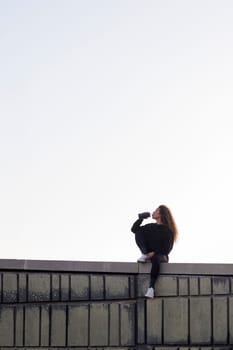 thirsty female athlete in sports clothes drinking beverage from bottle while resting on building roof under light sky, concept of urban sports and healthy lifestyle
