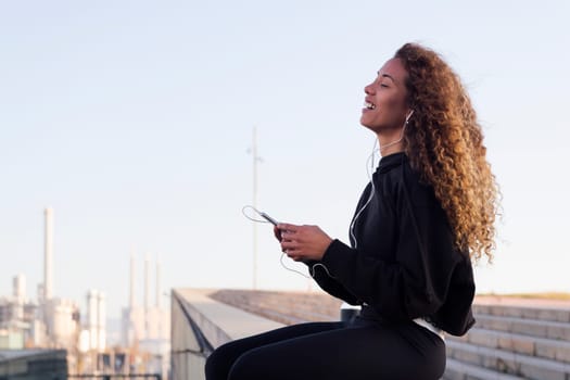 latin female athlete in sports clothes smiling happy while listening music on earphones from cellphone on building roof after workout, concept of urban sport and healthy lifestyle