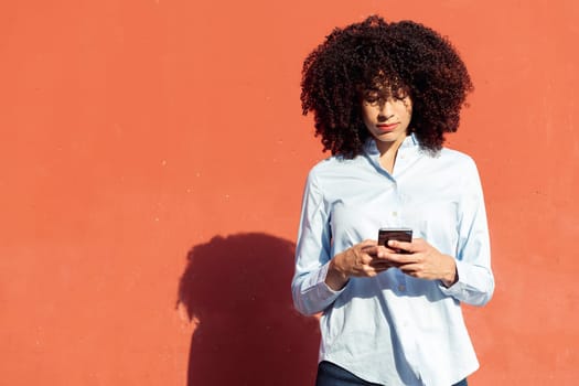 serious ethnic female entrepreneur in formal shirt texting on smartphone while standing in city on orange background