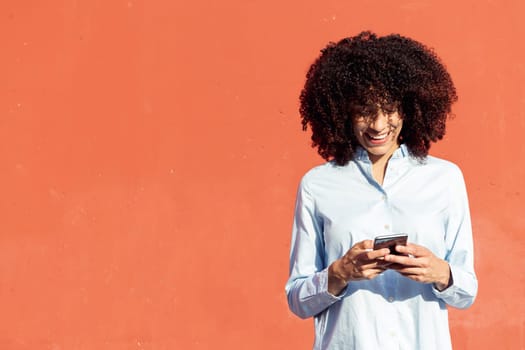 content ethnic female entrepreneur in formal shirt texting on smartphone while standing in city on orange background