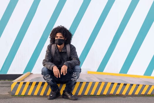 serious curly haired african american male teenager in black protective mask for flu prevention looking away thoughtfully sitting against striped wall on street