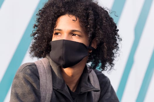 serious curly haired african american male teenager in black protective mask for flu prevention looking away thoughtfully against striped wall on street