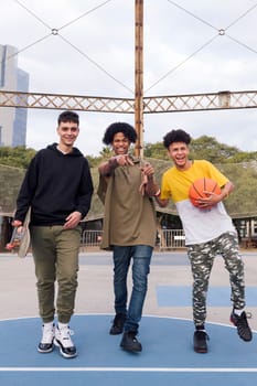 three confident multiracial male teens with basketball and skateboard walking joking and laughing happy while having fun in a sports court at park, concept of youth and urban lifestyle