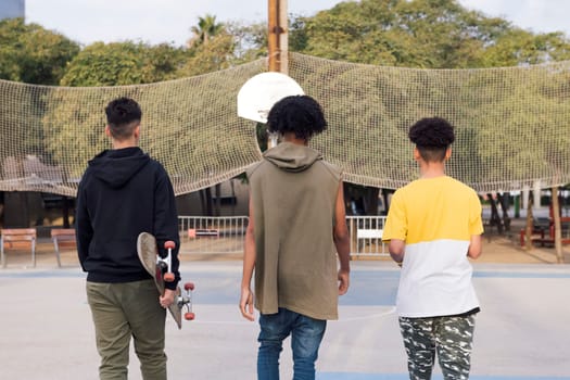 rear view of three unrecognizable multiracial male teens with skateboard walking in a sports court at park, concept of youth and urban lifestyle