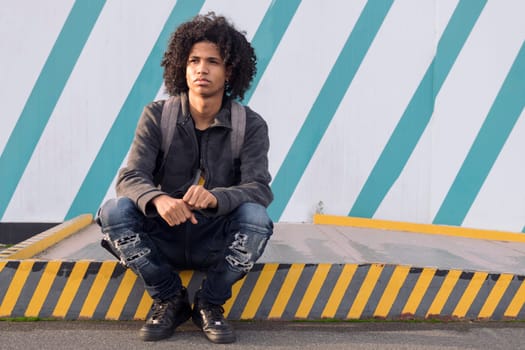 serious curly haired african american male teenager in informal street wear looking away thoughtfully sitting against striped wall on street