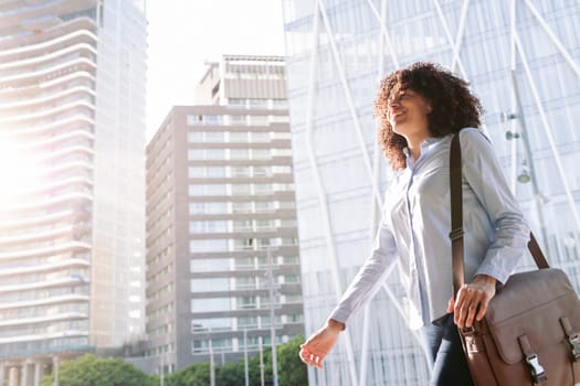 low angle side view of smiling female entrepreneur in smart casual outfit walking along street in city and looking away