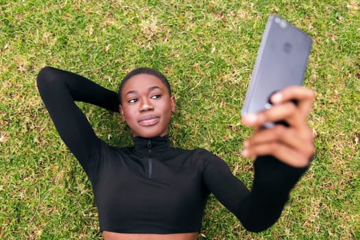young african american woman lying on the grass in a park smiling happy taking selfie photo on mobile phone, concept of technology and modern lifestyle