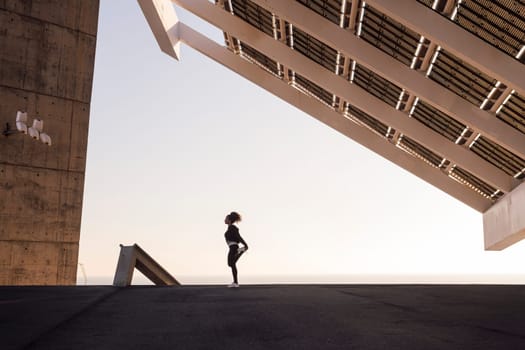 african american female athlete stretching during training under modern house while looking away in city, concept of urban sports and healthy lifestyle