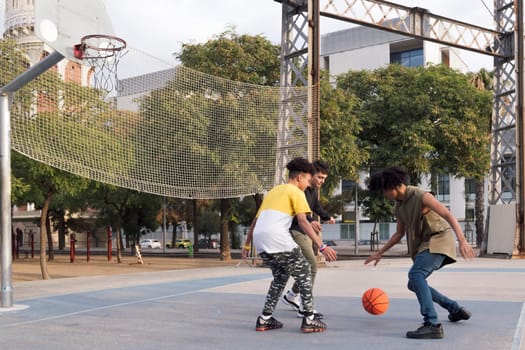 three focused multiethnic teen friends dribbling ball while playing basketball on sports ground in city park, concept of youth and urban lifestyle